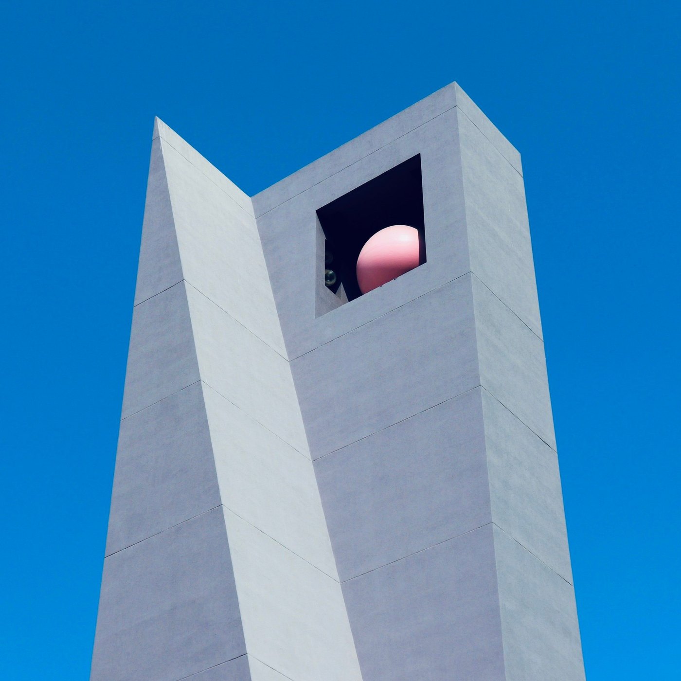 Pale concrete tower with a pink sphere in a square opening against a vivid blue sky