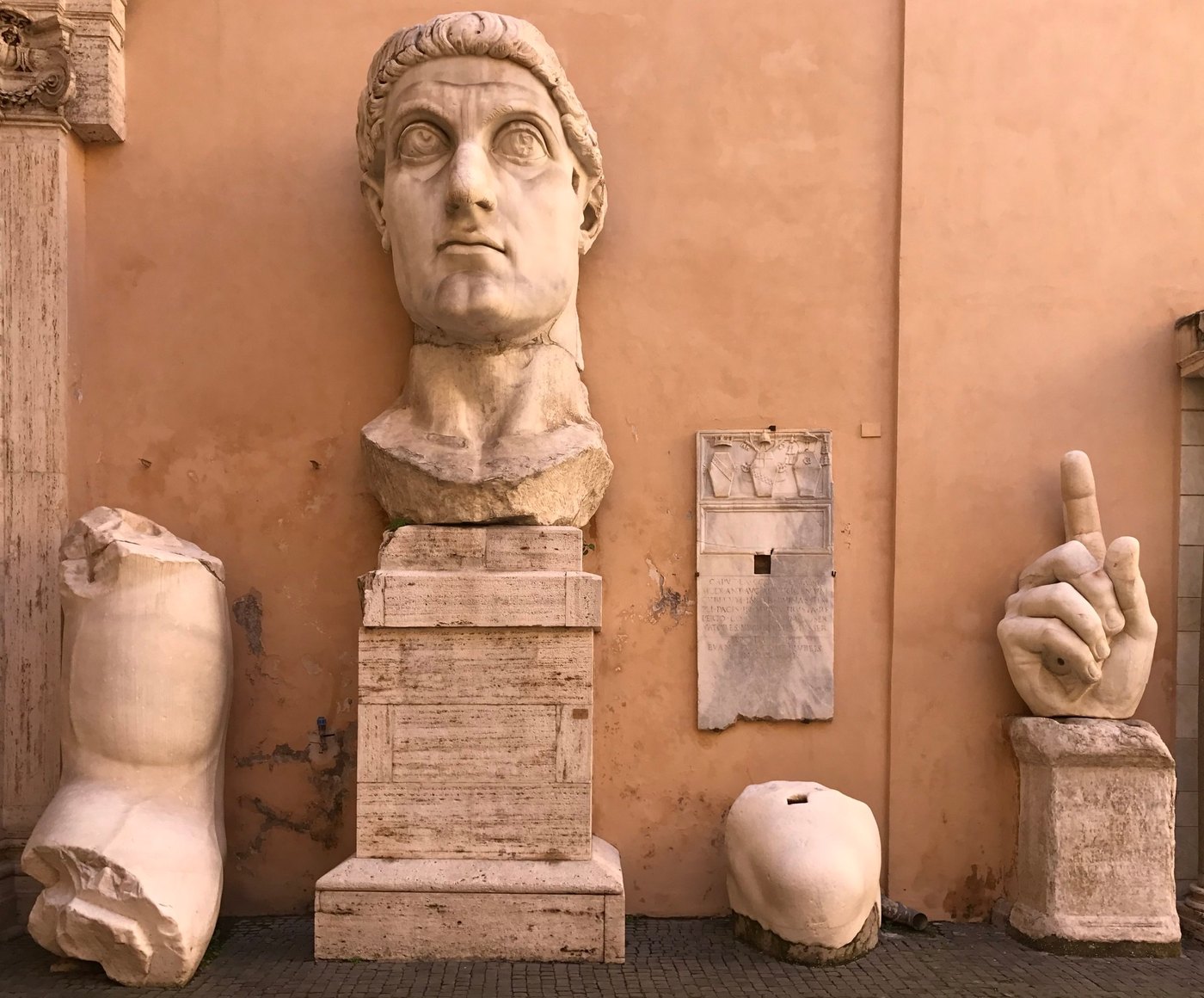 Colossal marble head, upraised marble hand and statue fragments along a peach wall