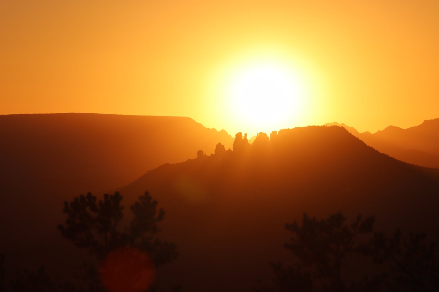 Sun blazing in an orange sky above silhouetted ridges, rock spires and dark pines