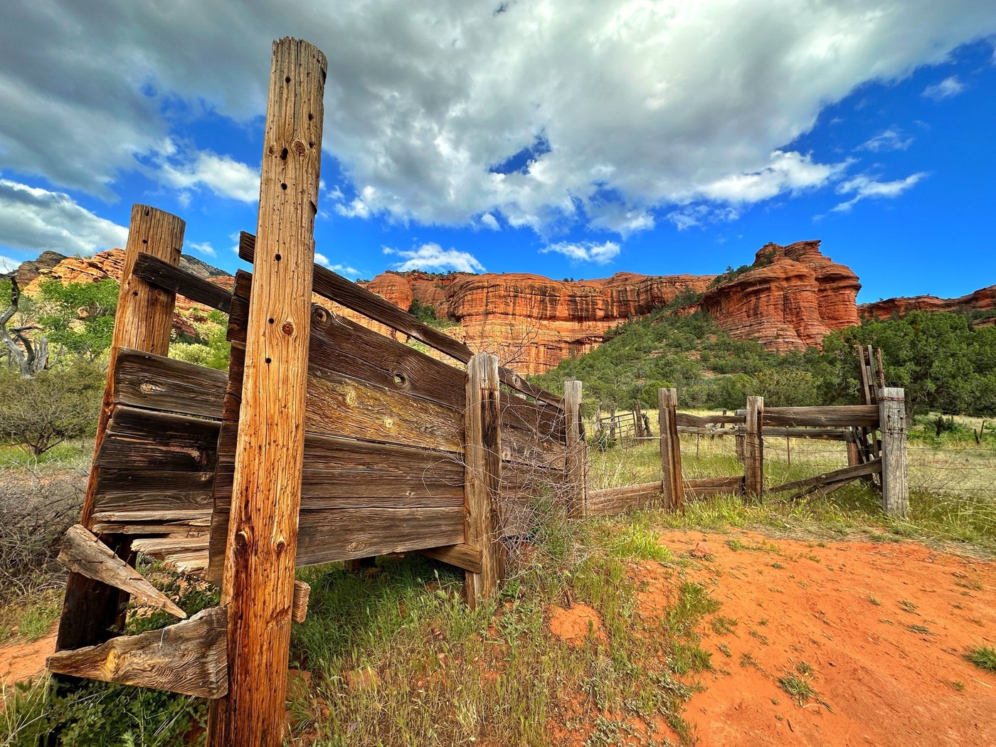Weathered wooden corral boards on red earth beneath red rock cliffs and a bright cloud-filled blue sky