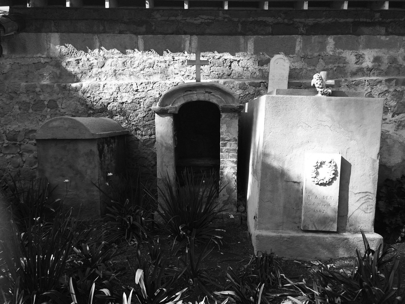 Black-and-white view of old stone tombs and a wreath plaque amid dark leafy plants