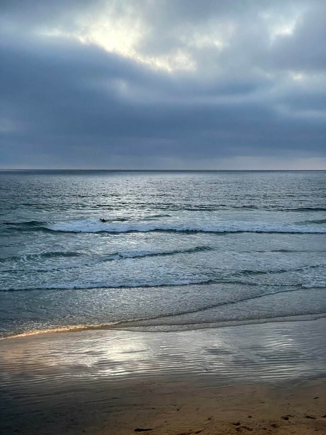 Overcast sky over gray ocean, with small waves and silvery light on wet sand